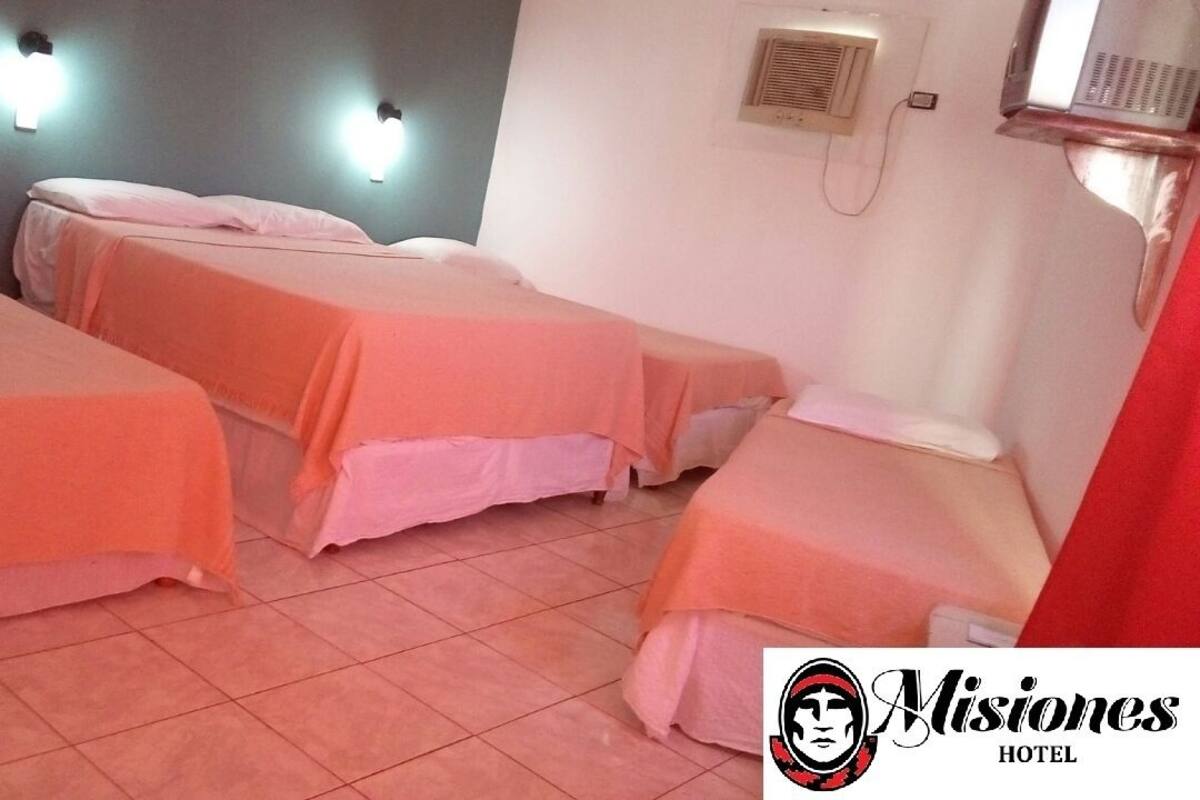 Habitación de hotel con tres camas individuales, cada una con una colcha rosa. Hay luces fijadas a la pared sobre cada cama y un calentador de pared a la derecha.