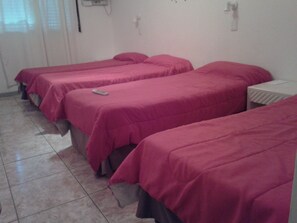 Habitación de hotel con tres camas, cada una con colcha rosa, una mesita de noche y un enchufe.