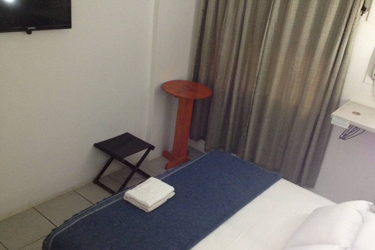 Habitación de hotel con cama, una mesita, una silla, televisión montada en la pared y una cortina.