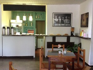 Un restaurante con mostrador verde, sillas de madera y una mesa con posavasos. Se observa un cuadro en la pared y una planta sobre la mesa.