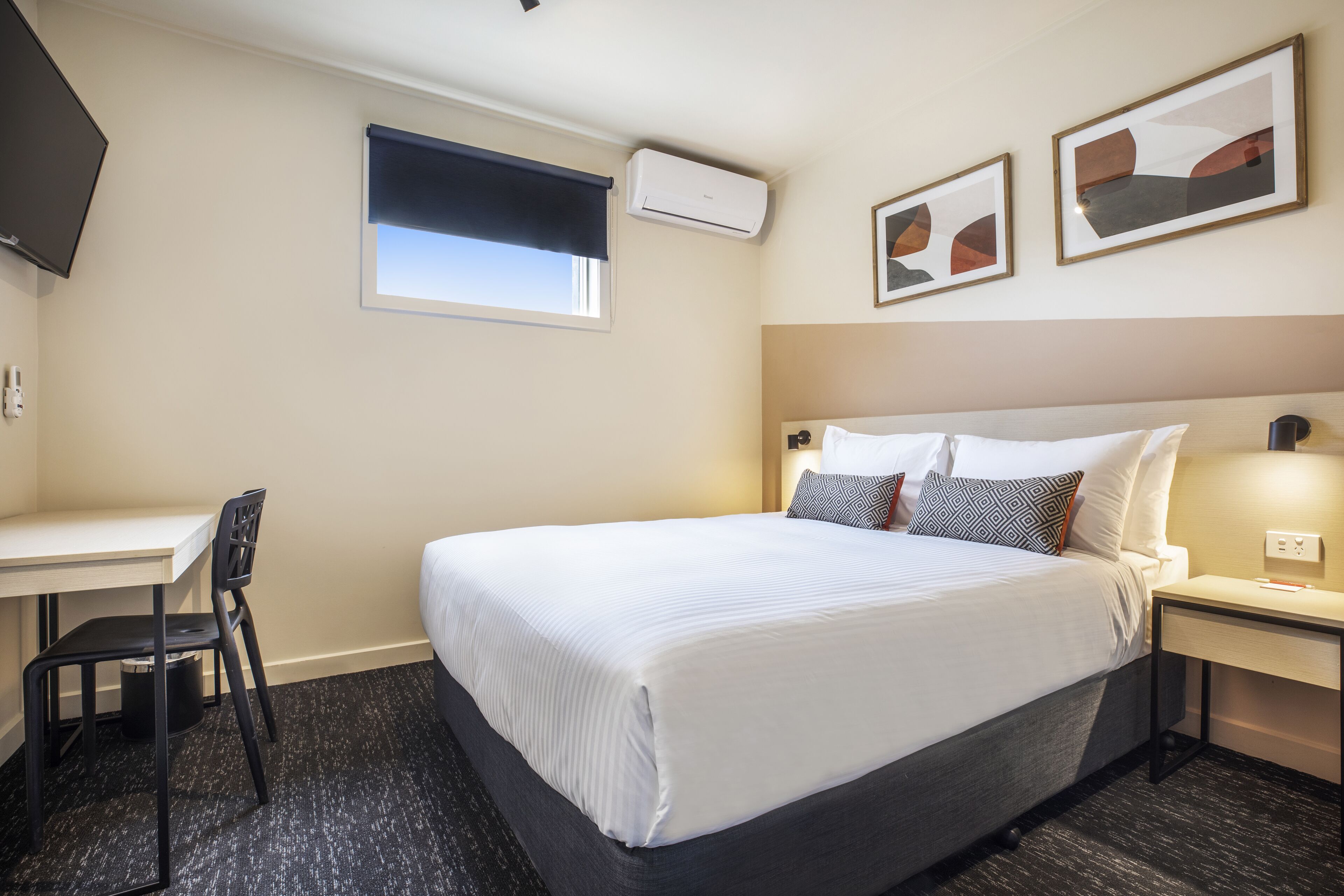 A hotel room with a bed, a desk, a chair, a window with a blind, and two framed pictures on the wall.