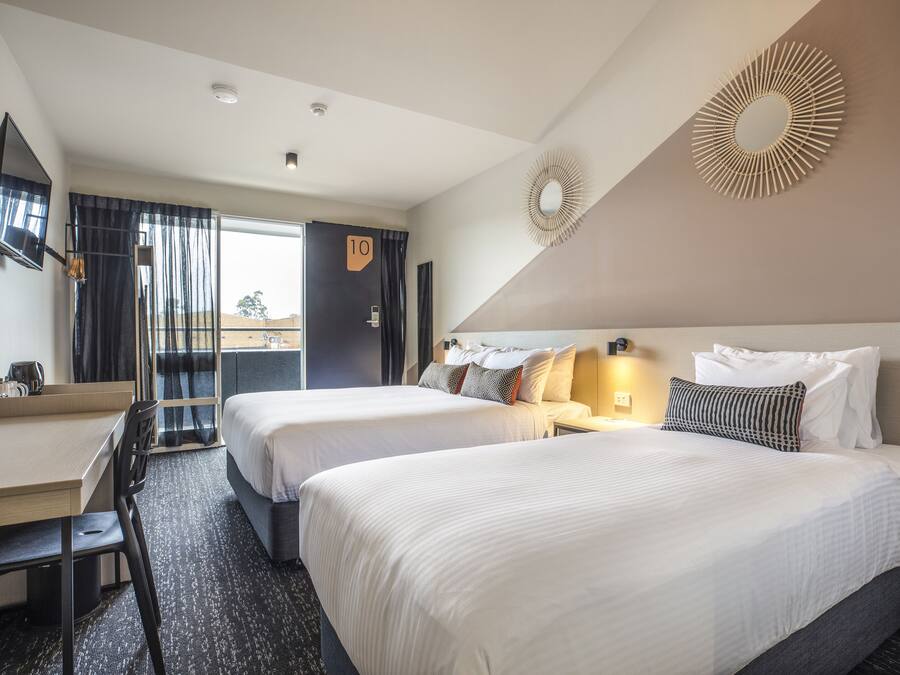 A hotel room with two beds, a desk, a chair, a TV, and a balcony with a view.