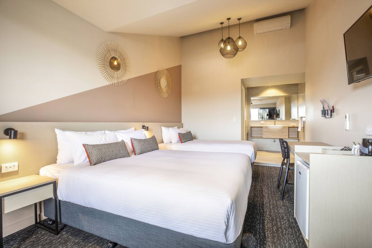 A hotel room with two beds, a desk, a chair, a small kitchenette, and a mirror.