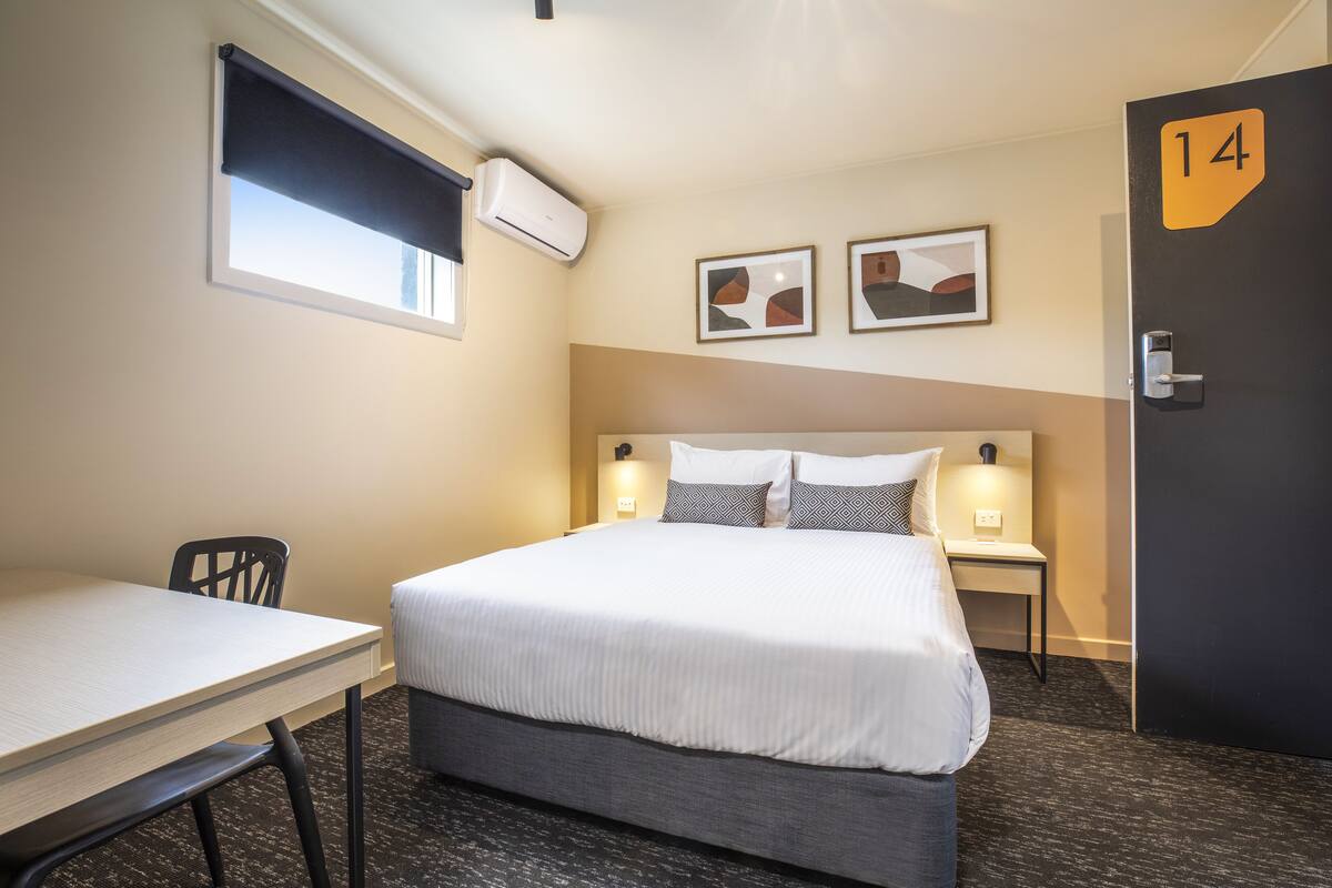 A hotel room with a bed, a desk, a chair, two wall-mounted artworks, an air conditioning unit, and a door marked with the number 14.