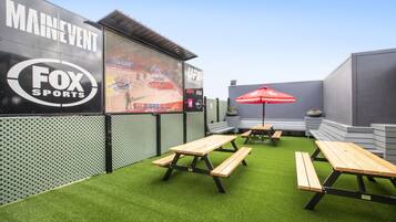 An outdoor seating area with picnic tables and a red umbrella, surrounded by artificial grass and a wall with advertisements.