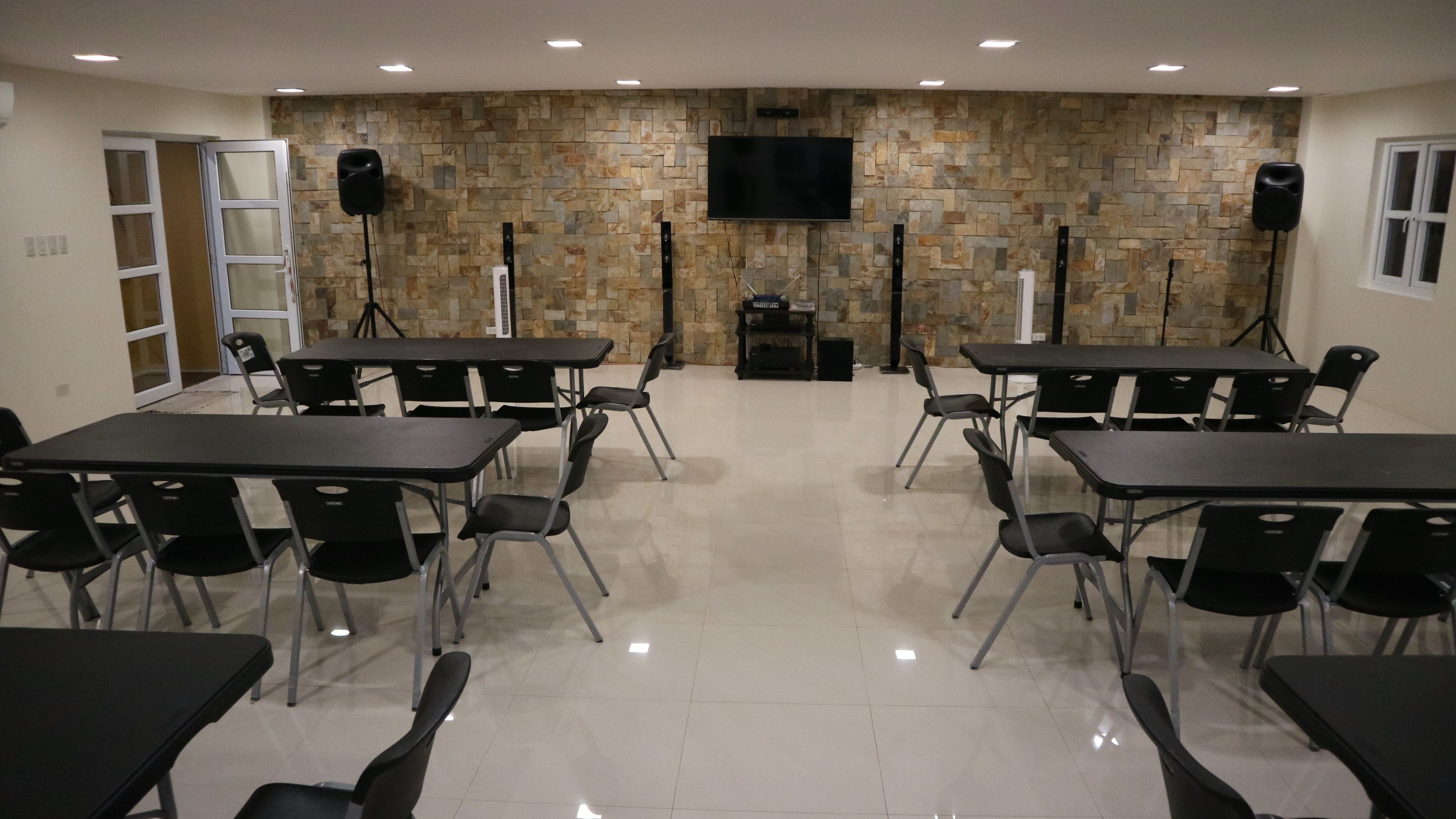 Una sala conferenze con diversi tavoli e sedie, una parete in pietra, una TV a parete e degli altoparlanti.