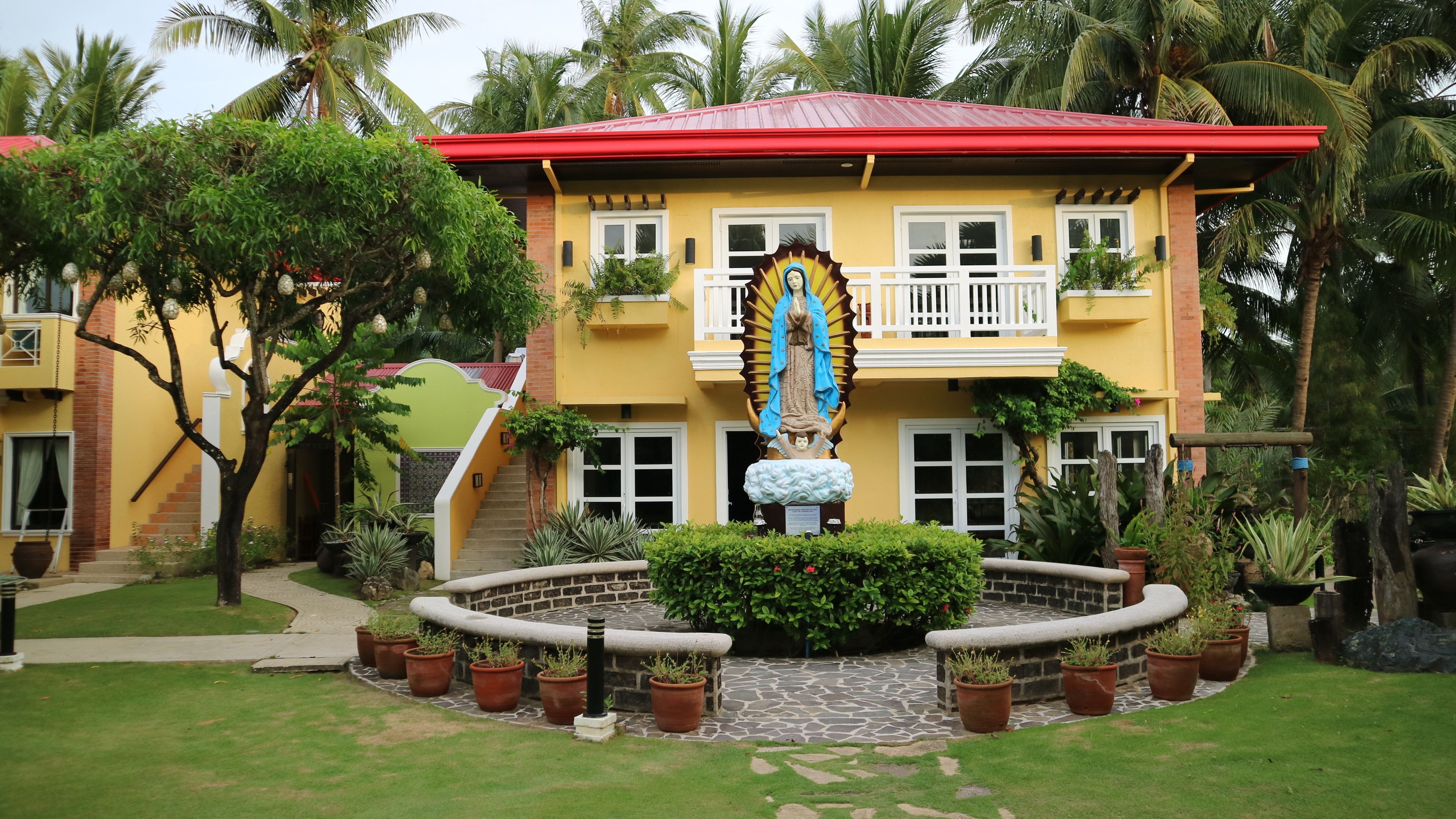 Un edificio de dos pisos con techo rojo, una estatua de una figura religiosa y un jardín bien cuidado con plantas en macetas.