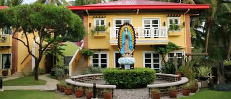 Un edificio de dos pisos con techo rojo, una estatua de una figura religiosa y un jardín bien cuidado con plantas en macetas.