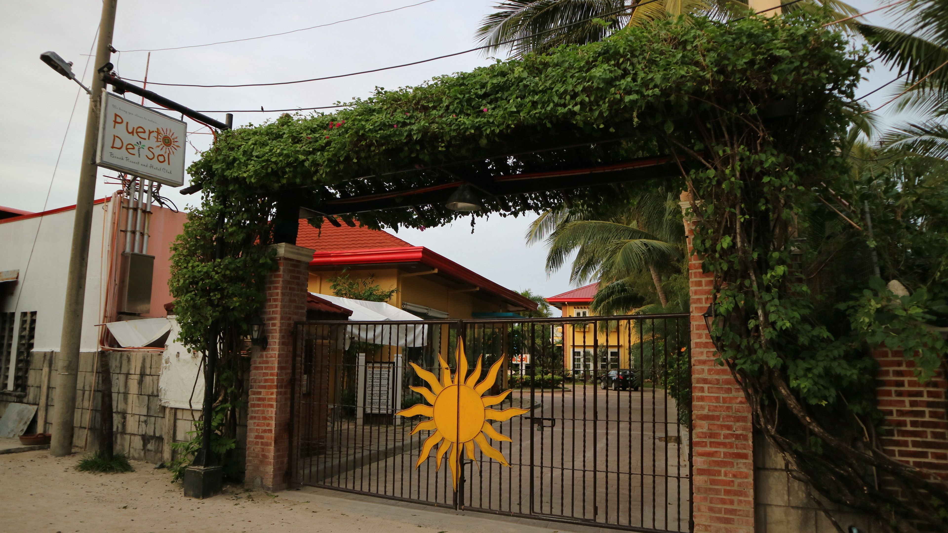 A gated entrance with a sun symbol, a sign for Puerto del Sol, and a building with a red roof.