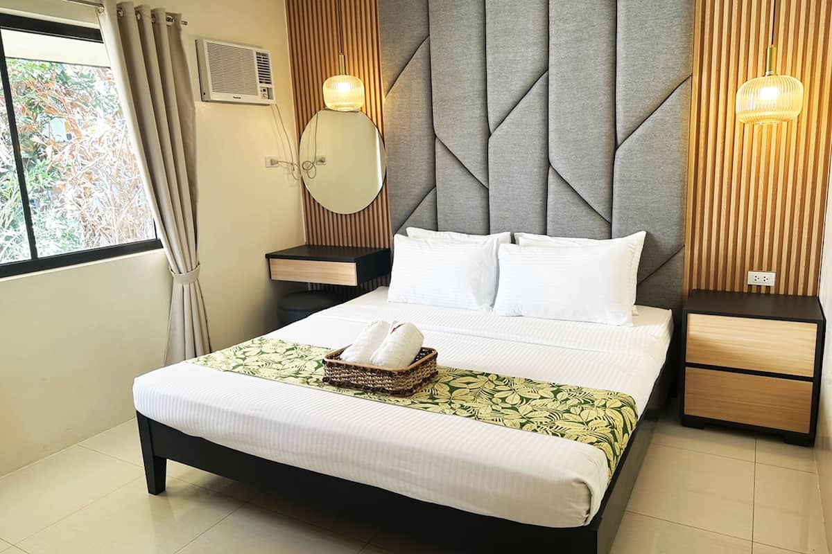 A neatly made bed with white linens, a bedside table with a lamp, and a window with curtains.