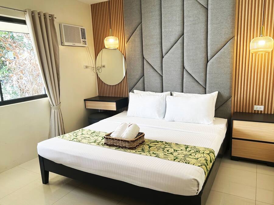 Una cama bien hecha con sábanas blancas, una mesita de noche con lámpara y una ventana con cortinas.