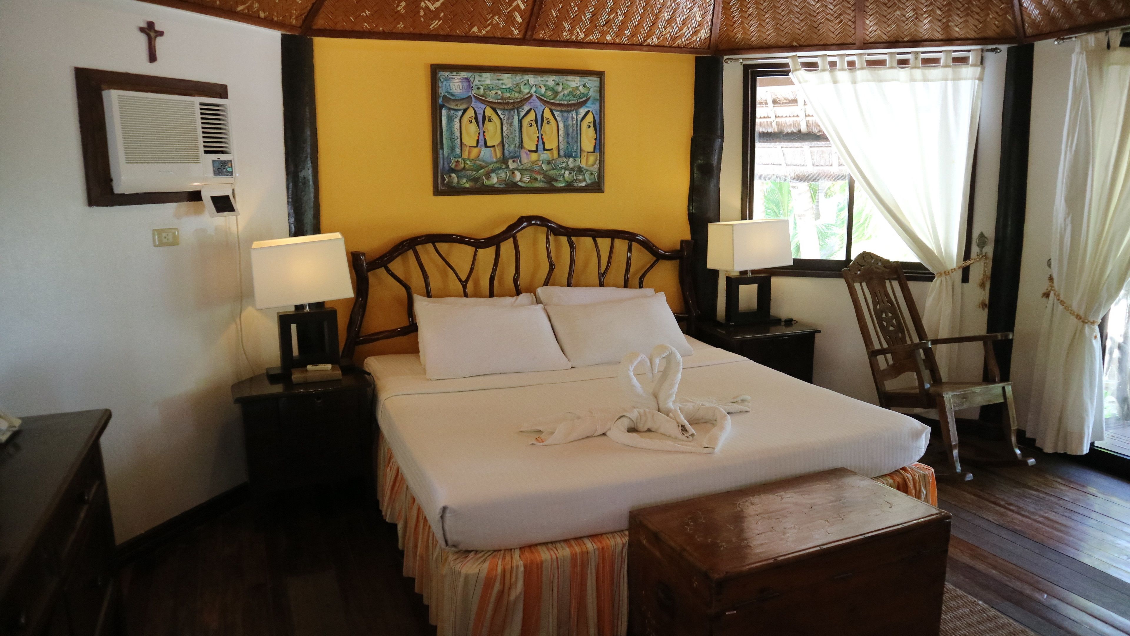 A hotel room with a bed, two bedside tables with lamps, a wooden headboard, a painting on the wall, a cross on the wall, and a window with curtains.
