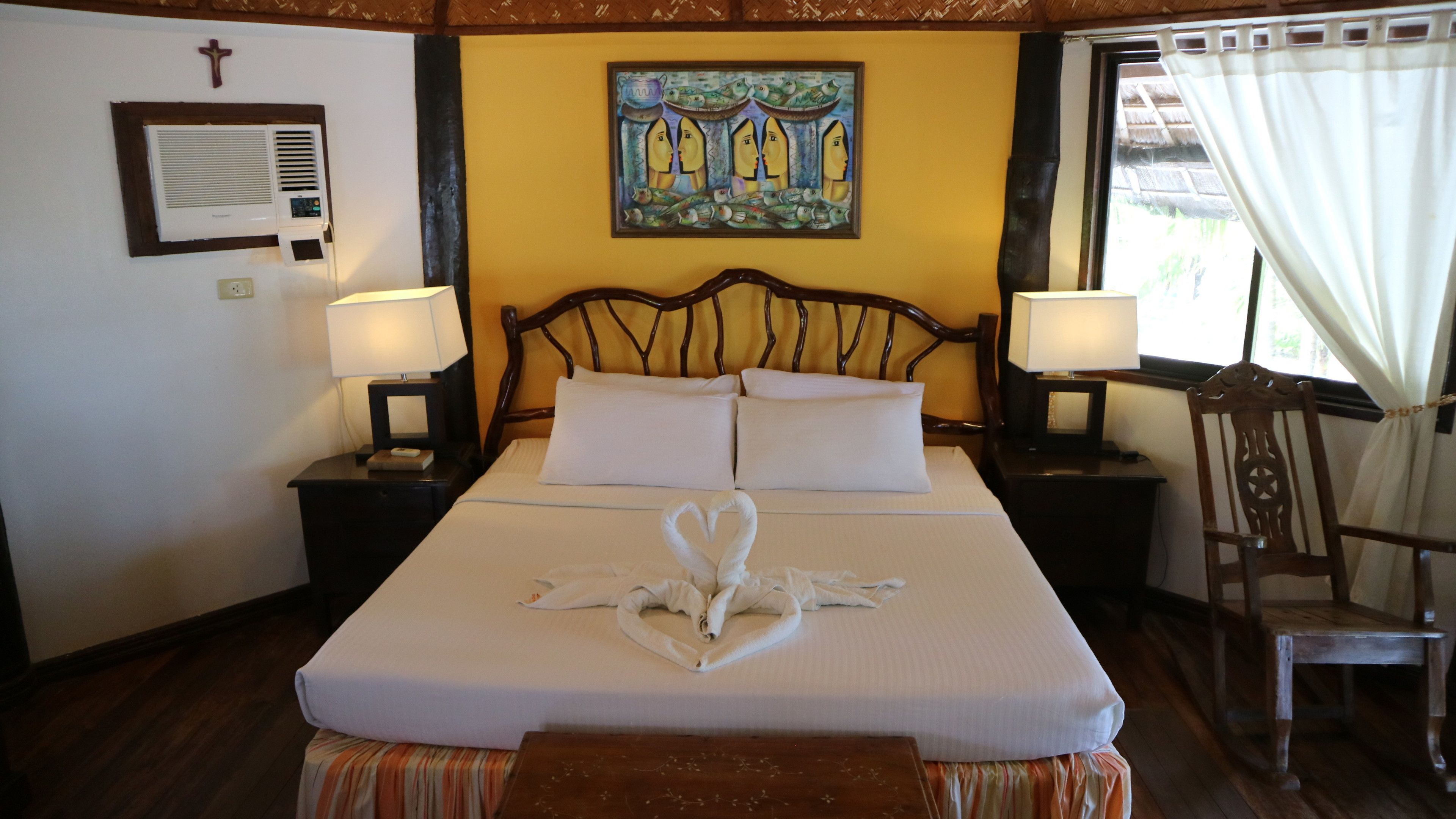 A neatly made bed with white linens and a heart-shaped towel arrangement, a wooden headboard, two bedside lamps, a wall-mounted air conditioner, and a painting above the bed.