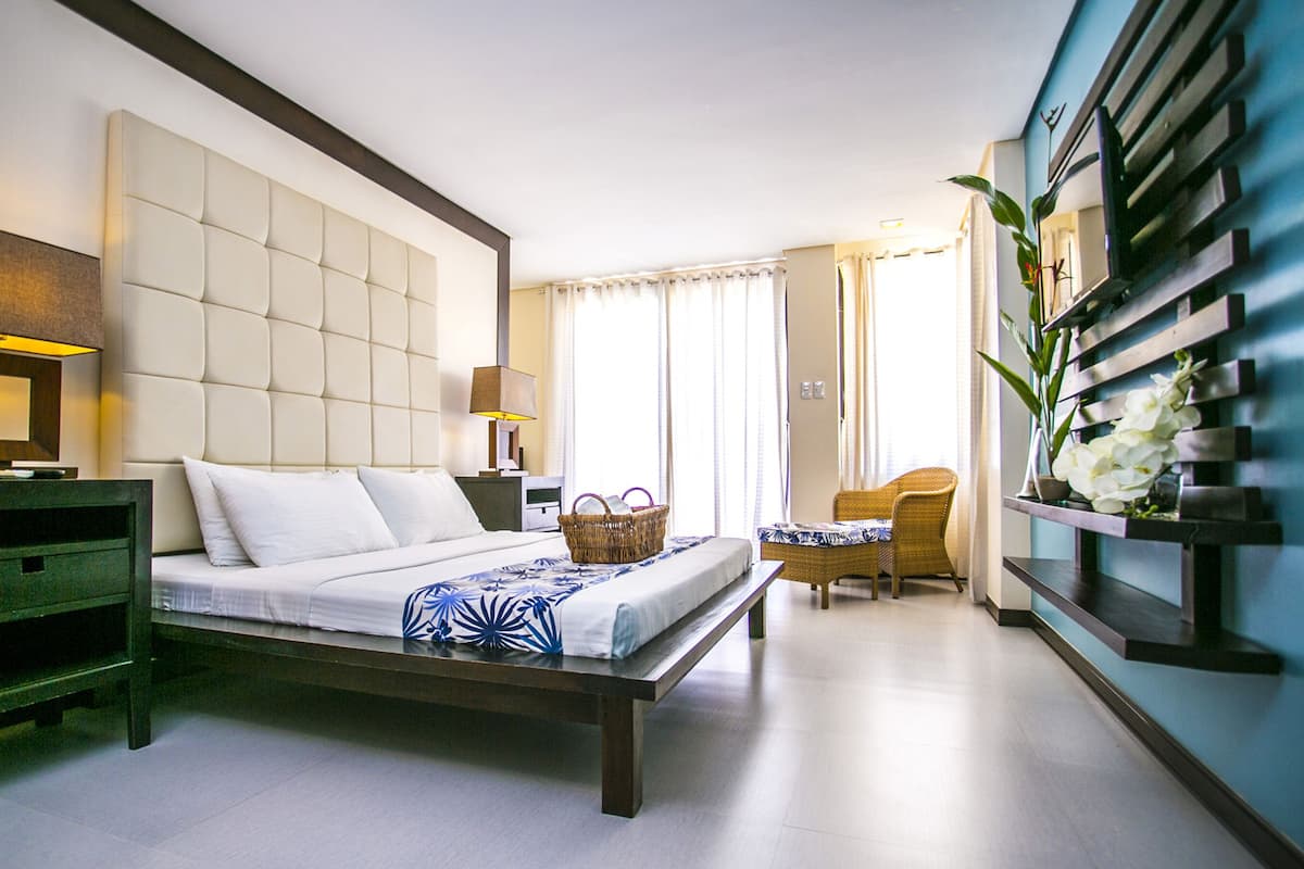 A hotel room with a large bed, a bedside table with a lamp, a chair, a small table with a basket, and a wall-mounted shelf with flowers.