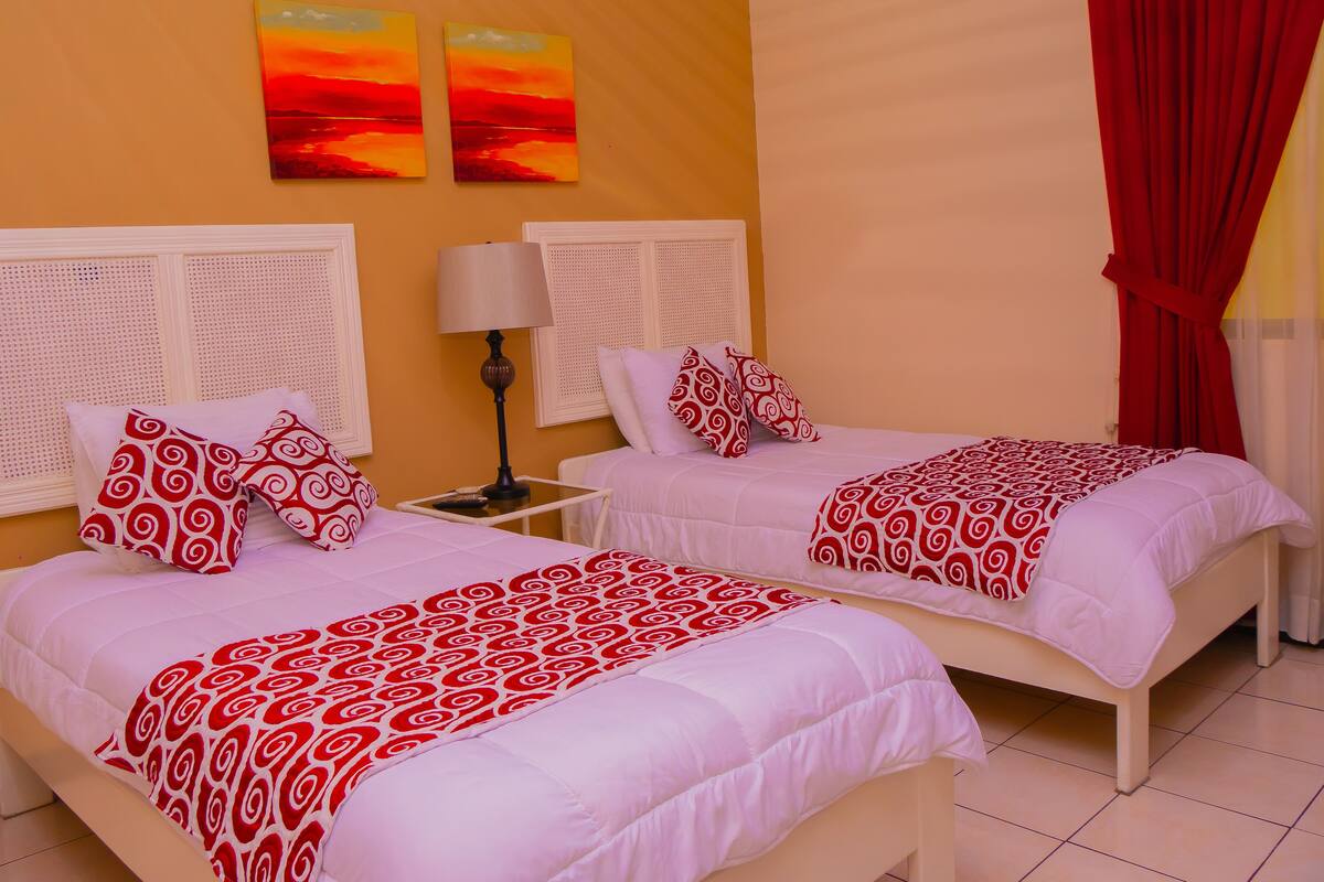 Ein Hotelzimmer mit zwei Betten, jeweils mit weißer Bettwäsche und Kissen in Rot-Weiß-Muster. An den Wänden hängen zwei Bilder, die Sonnenuntergänge zeigen, eine weiße Lampe steht auf einem Nachttisch und ein roter Vorhang ist am Fenster.
