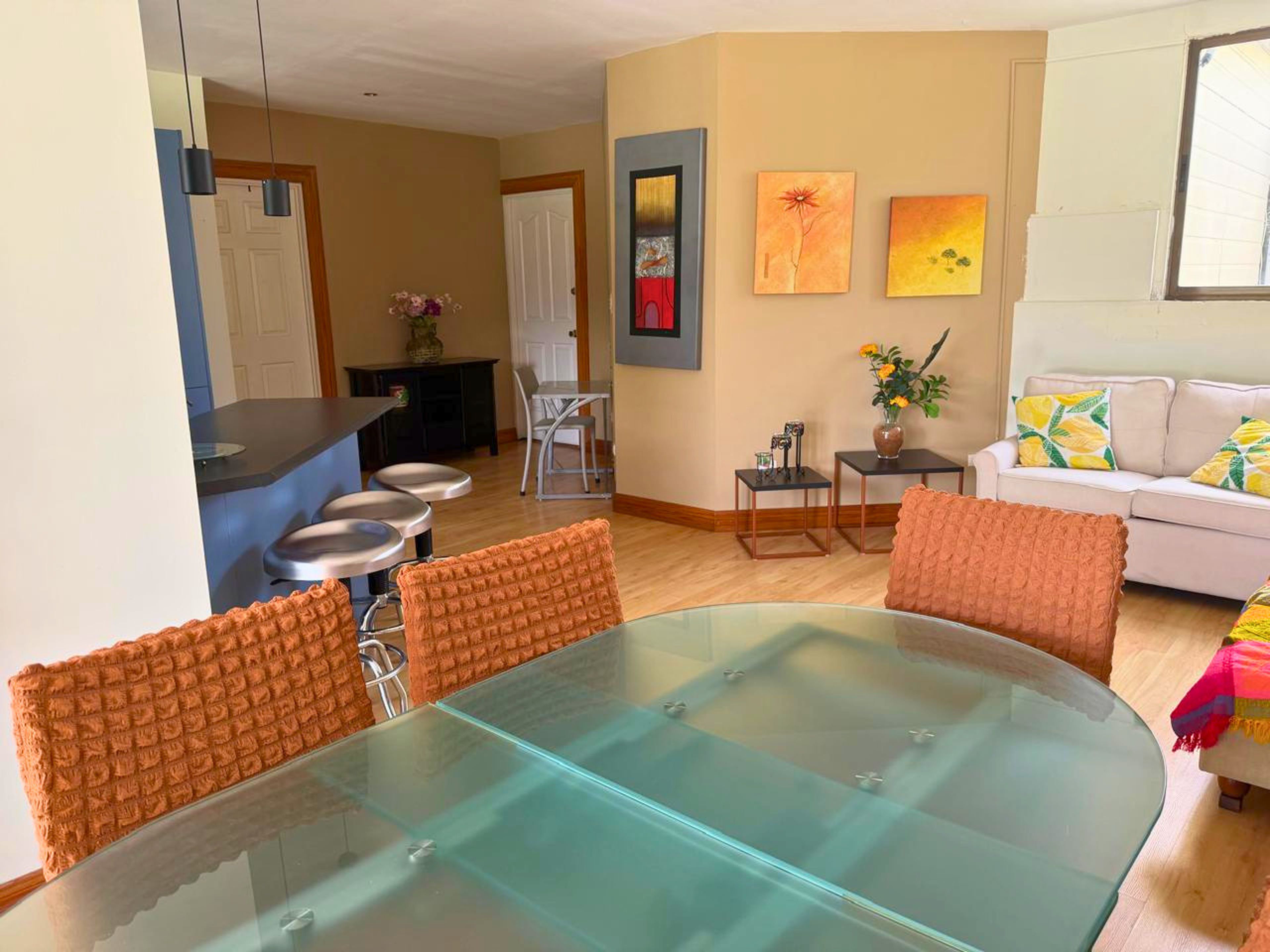 Una sala de estar moderna con una mesa de comedor de vidrio, sillas con cojines naranja, un sofá blanco y una pequeña mesa auxiliar con un jarrón.