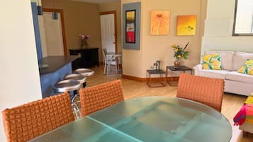 Una sala de estar moderna con una mesa de comedor de vidrio, sillas con cojines naranja, un sofá blanco y una pequeña mesa auxiliar con un jarrón.