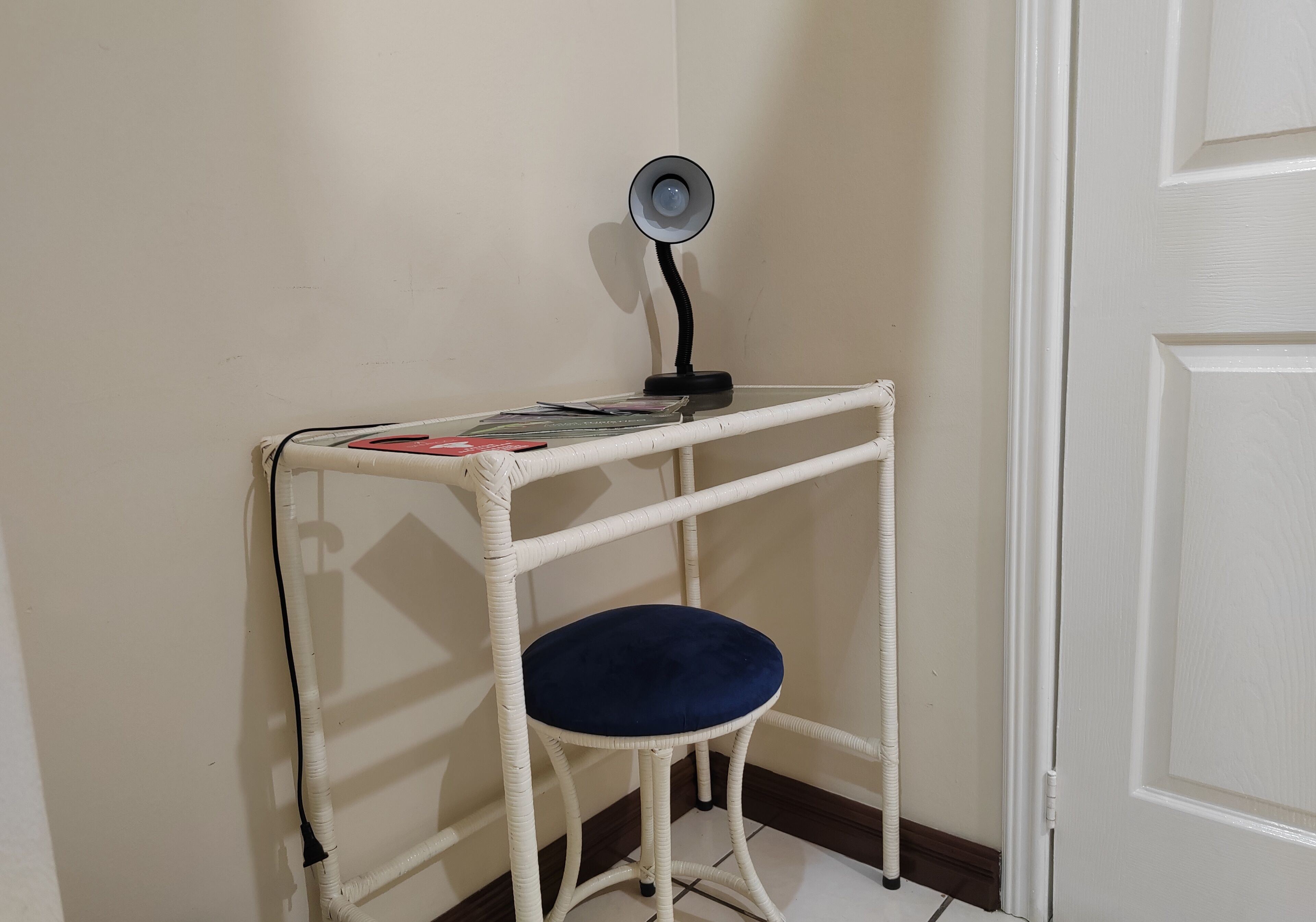 A corner room with a white door, a white wicker desk with a blue cushioned seat, and a small table with books and a lamp.