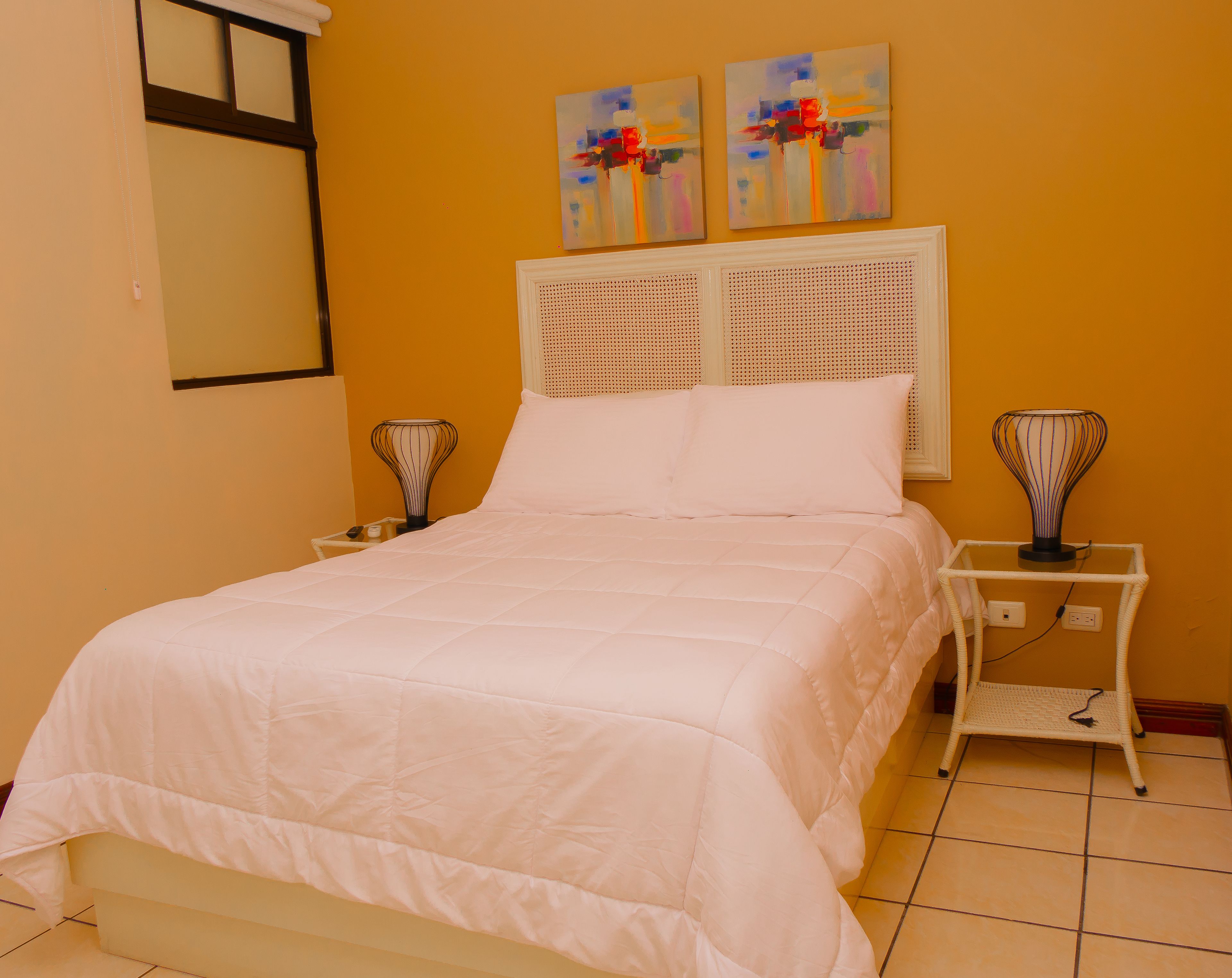 Un dormitorio con una cama, dos mesitas de noche, un tocador, un espejo montado en la pared y dos cuadros abstractos.