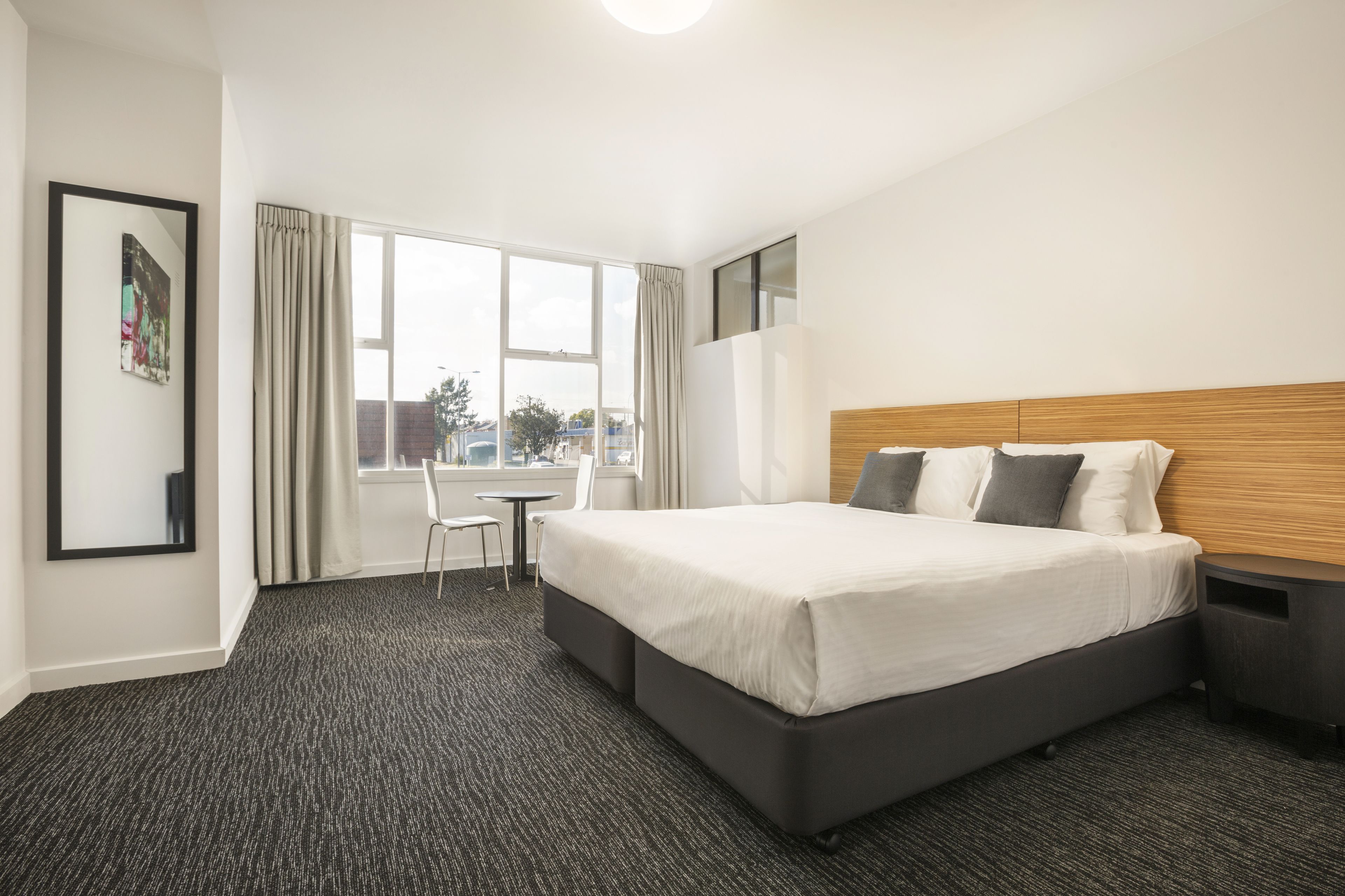 A hotel room with a large bed, a desk, and a chair. There is a window with curtains, a picture on the wall, and a view of the city.