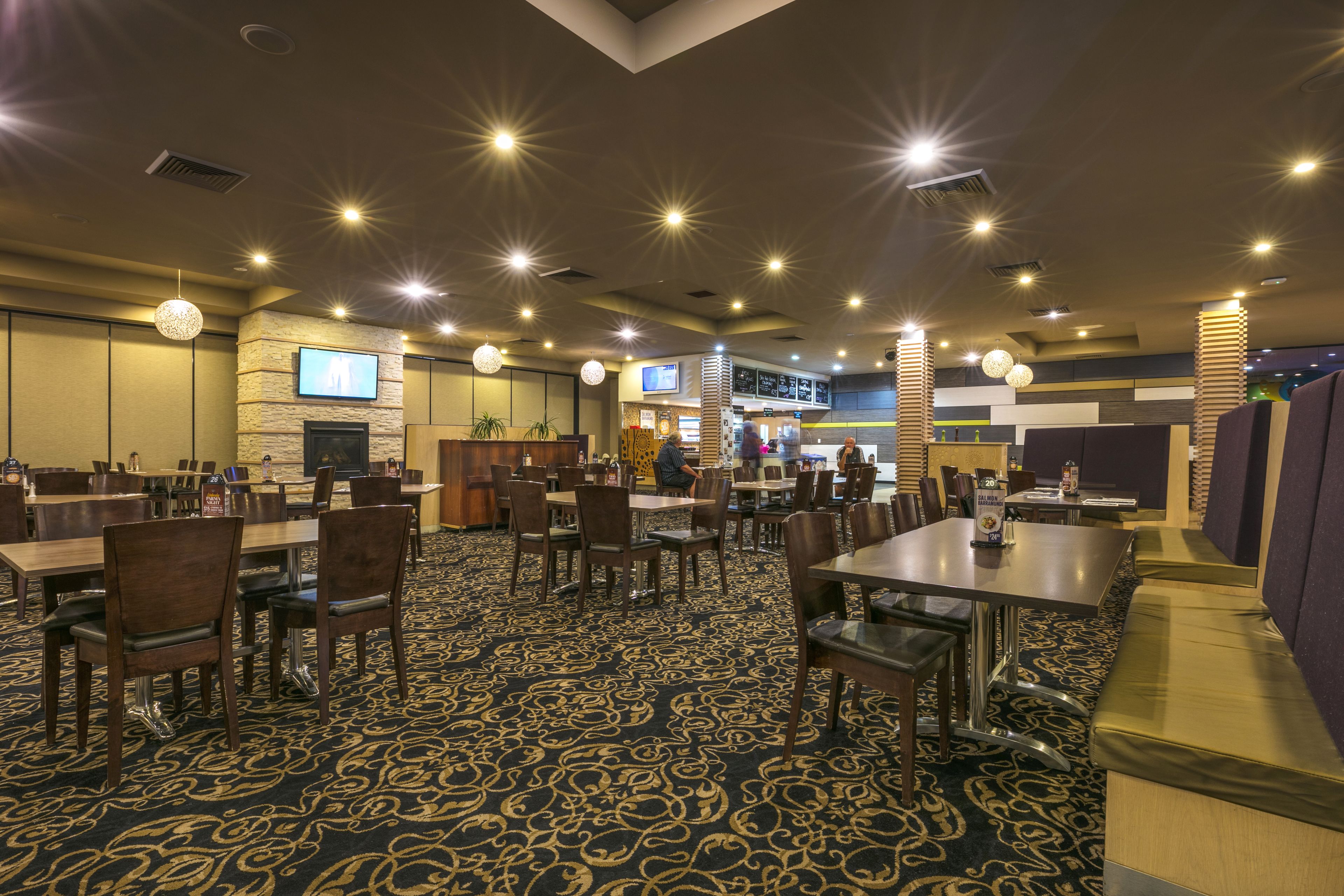 A hotel lobby with multiple dining tables and chairs, a large screen on the wall, and a patterned carpet.