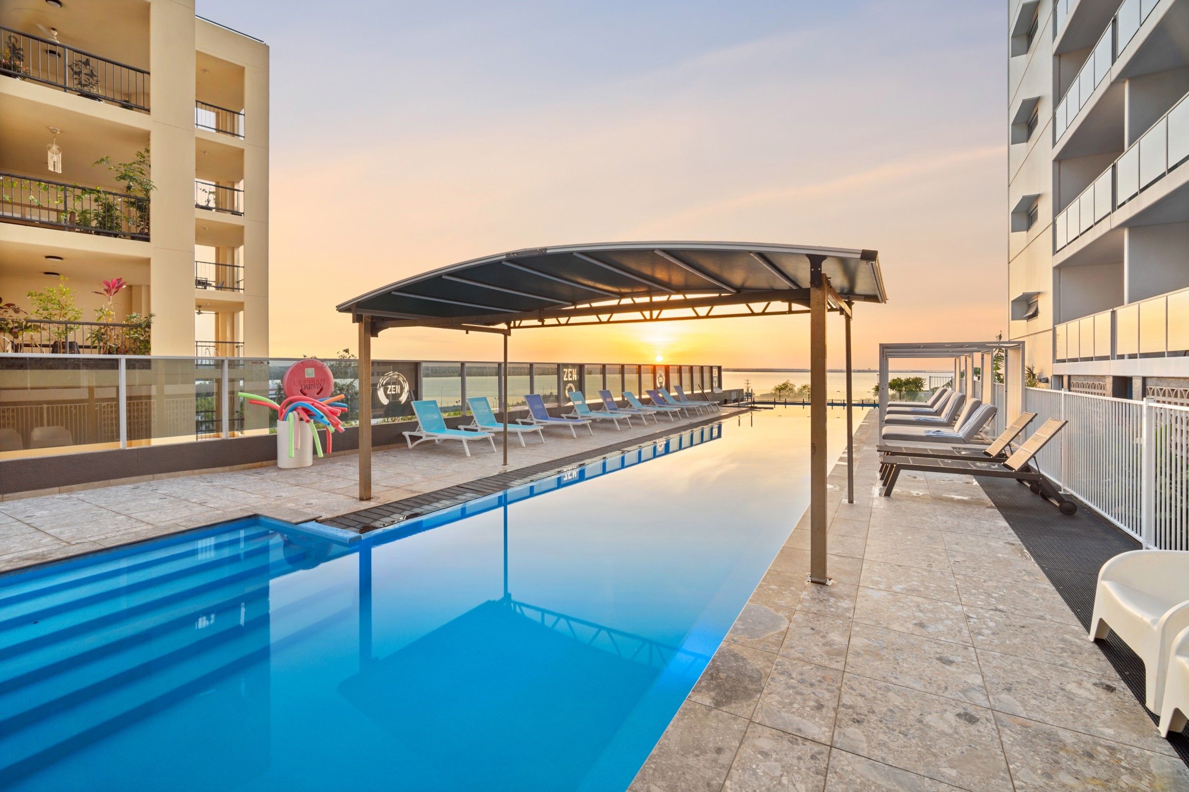 Piscina con sillas de descanso y zona de estar cubierta, rodeada por un edificio con balcones.