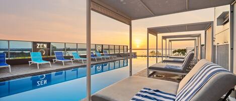A rooftop pool area with lounge chairs and a covered seating area, overlooking a sunset view.