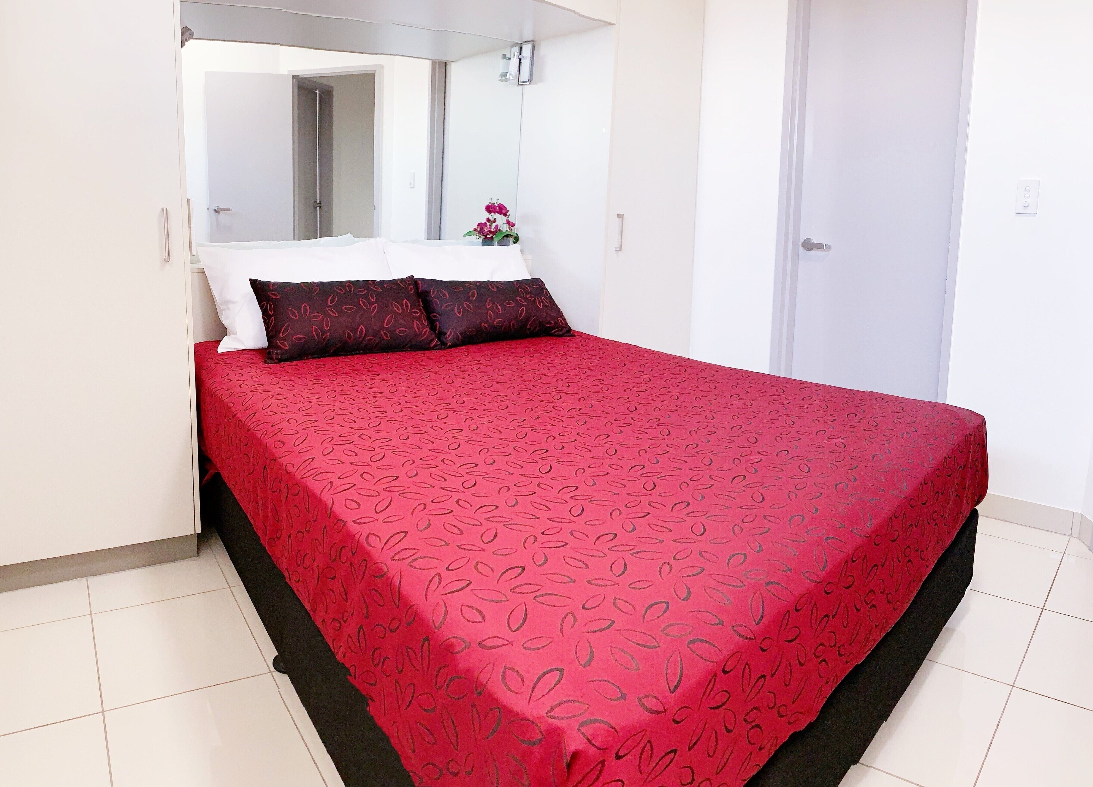 A neatly made bed with a red patterned bedspread and two pillows, a white wall, and a door in the background.