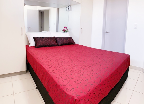 A neatly made bed with a red patterned bedspread and two pillows, a white wall, and a door in the background. - Ramada Suites by Wyndham Zen Quarter Darwin (Darwin)