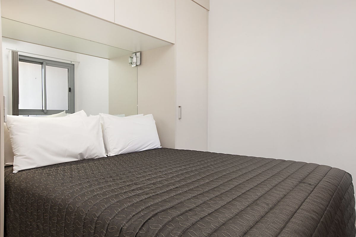 Un lit bien fait, recouvert d’une couvre-lit à motifs sombres et agrémenté d’oreillers blancs, un grand miroir au-dessus du lit et une fenêtre dont les stores sont fermés.