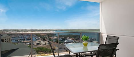A balcony with a glass table and chairs, overlooking a marina and cityscape.
