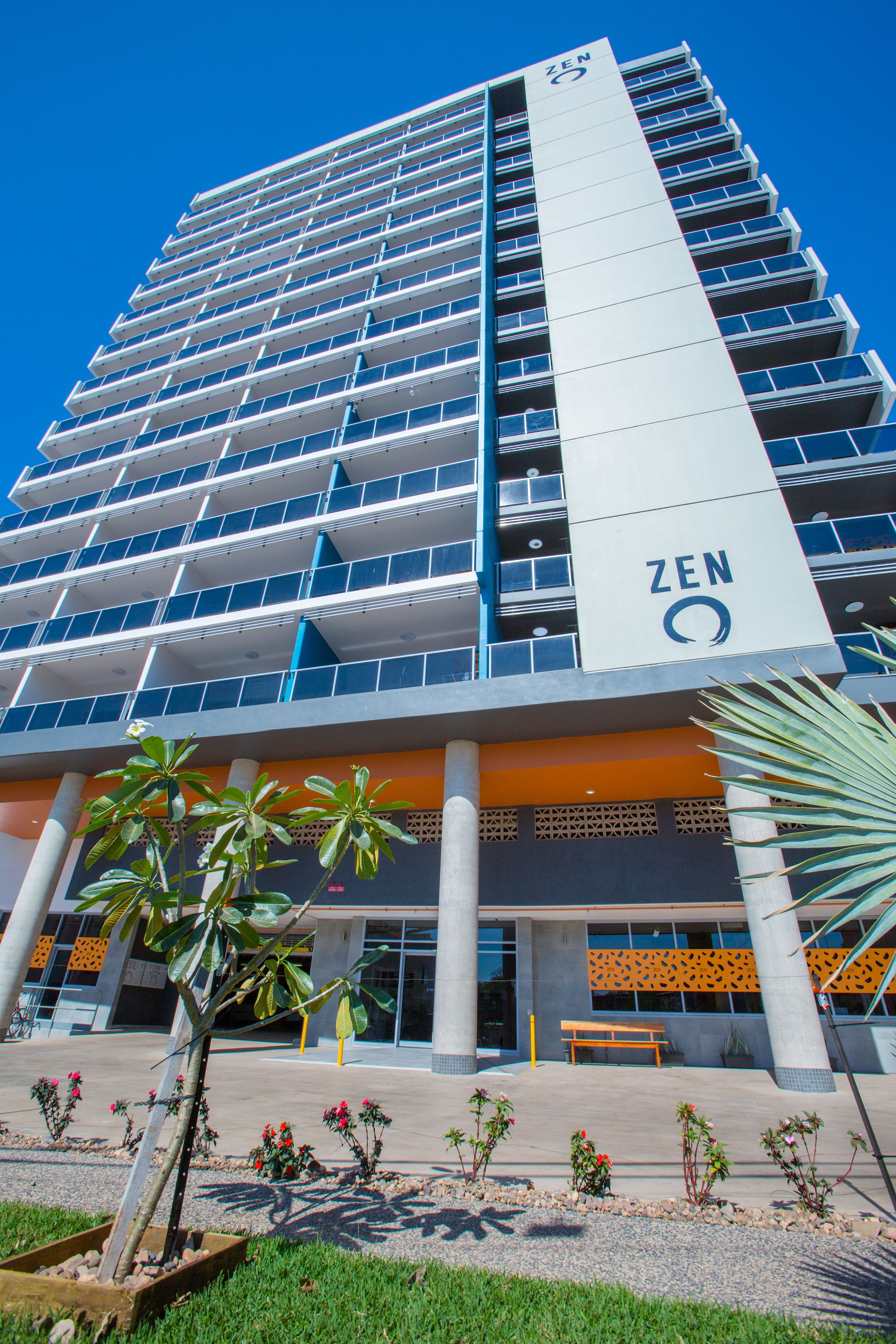 Un edificio moderno con un llamativo letrero 'ZEN', que presenta una fachada estructurada con varios balcones y una zona de entrada central con un banco y palmeras.