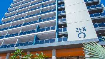 Un edificio moderno con un llamativo letrero 'ZEN', que presenta una fachada estructurada con varios balcones y una zona de entrada central con un banco y palmeras.