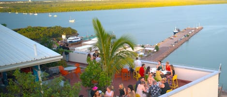 Un bar en la azotea con mesas al aire libre que da a un cuerpo de agua, un muelle con barcos y vistas del paisaje circundante.