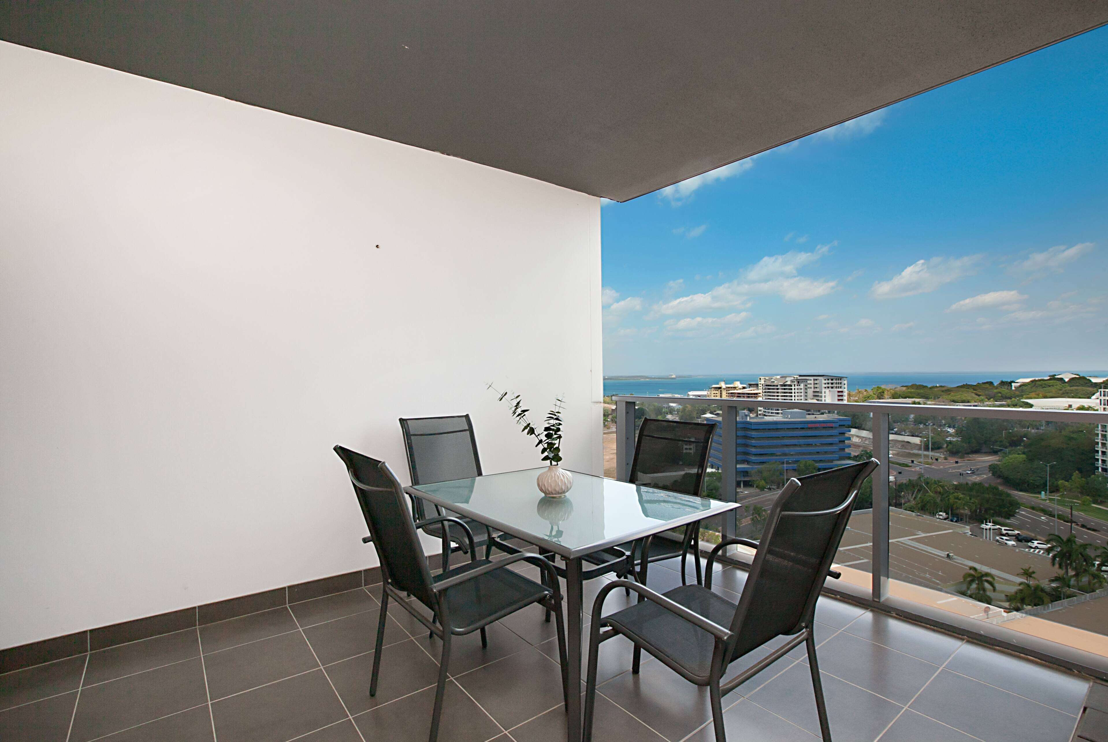 A balcony with a glass table and chairs, a potted plant, and a city view.