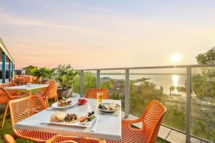 Un área de comedor al aire libre con mesas y sillas, vista a un cuerpo de agua y un atardecer de fondo.