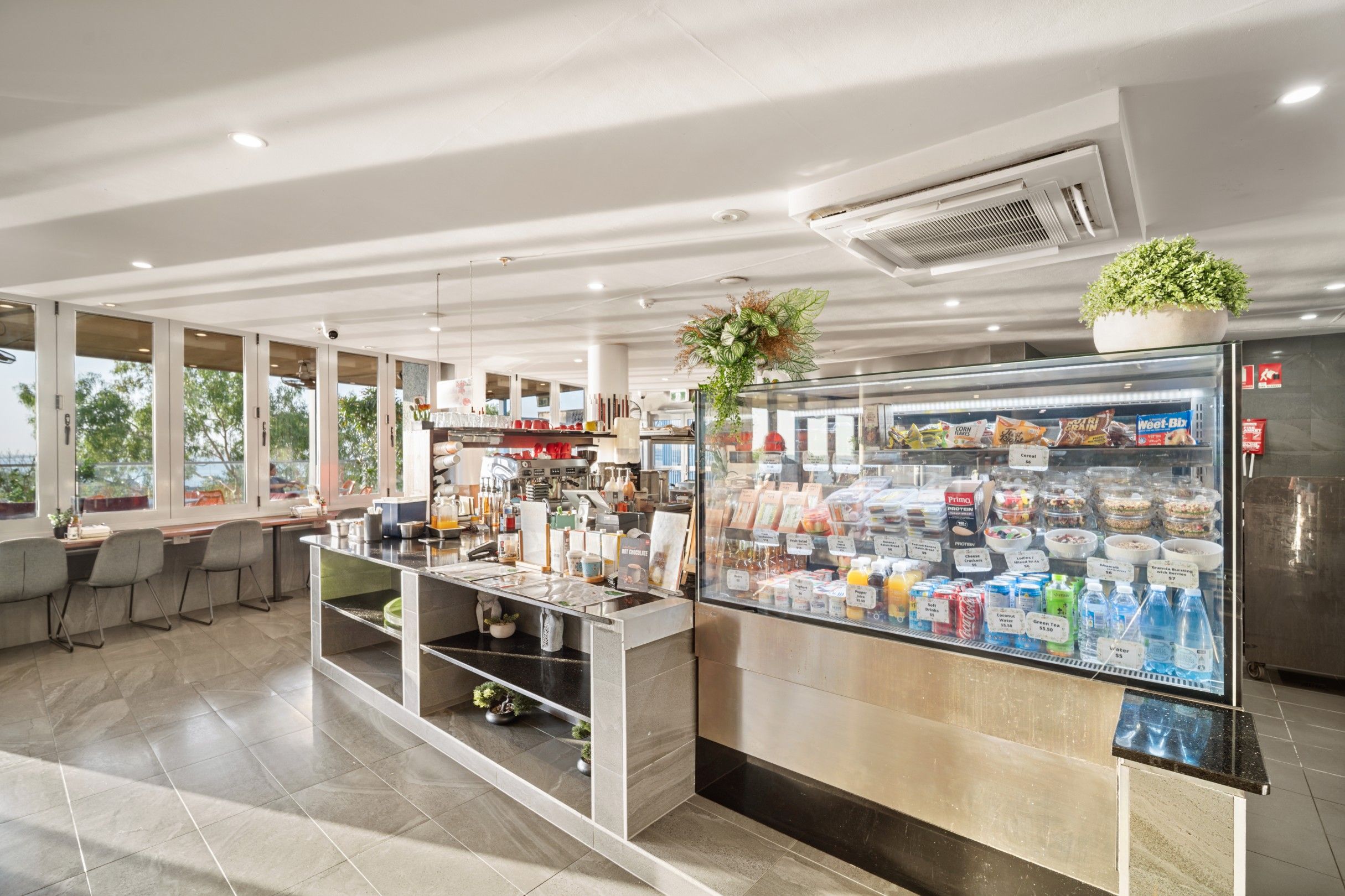 A modern cafe with a self-service counter, a refrigerated display case with snacks, and a seating area with tables and chairs.
