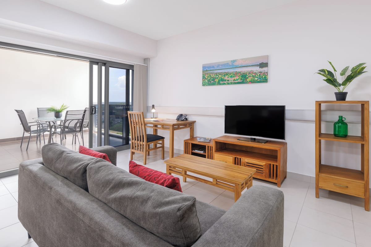 Un salon moderne avec un canapé gris, une table basse en bois et un meuble pour la télévision. Il y a un balcon avec une table et des chaises, ainsi qu'un tableau accroché au mur.