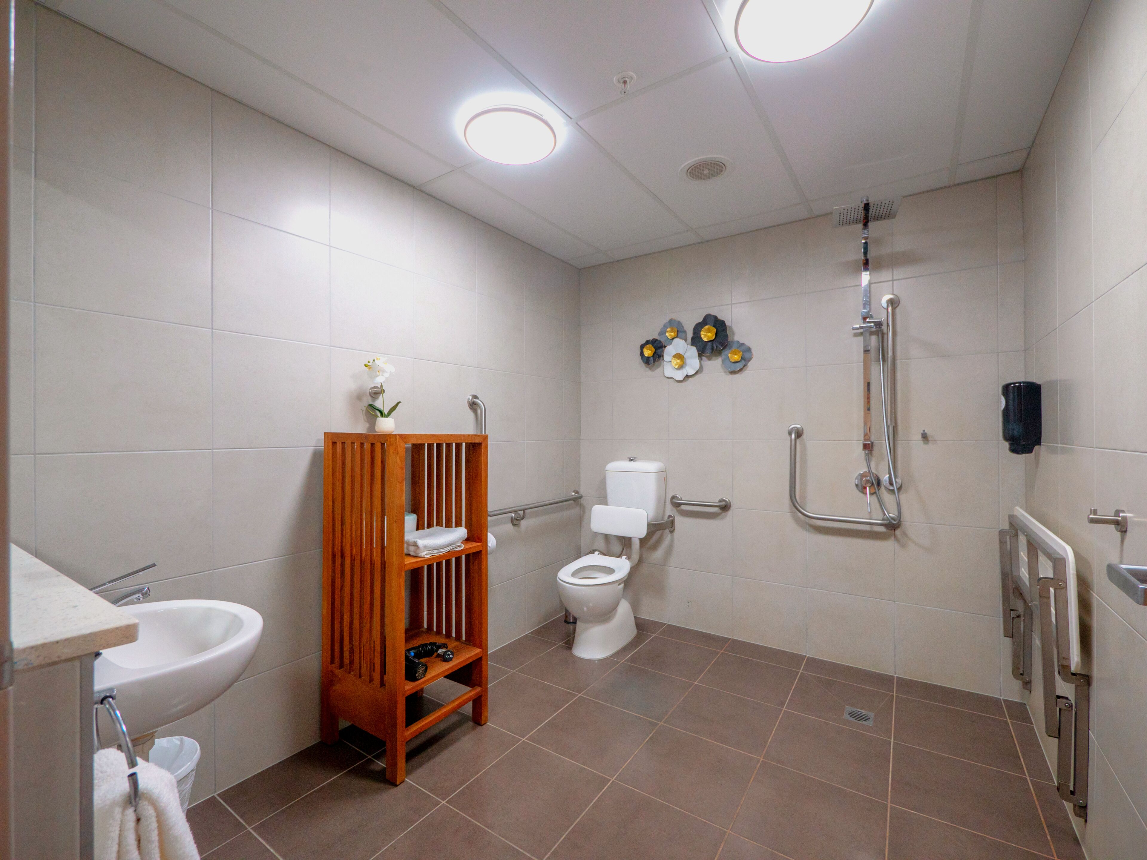 A bathroom with a sink, toilet, and shower. It includes a wooden shelf and handrails.