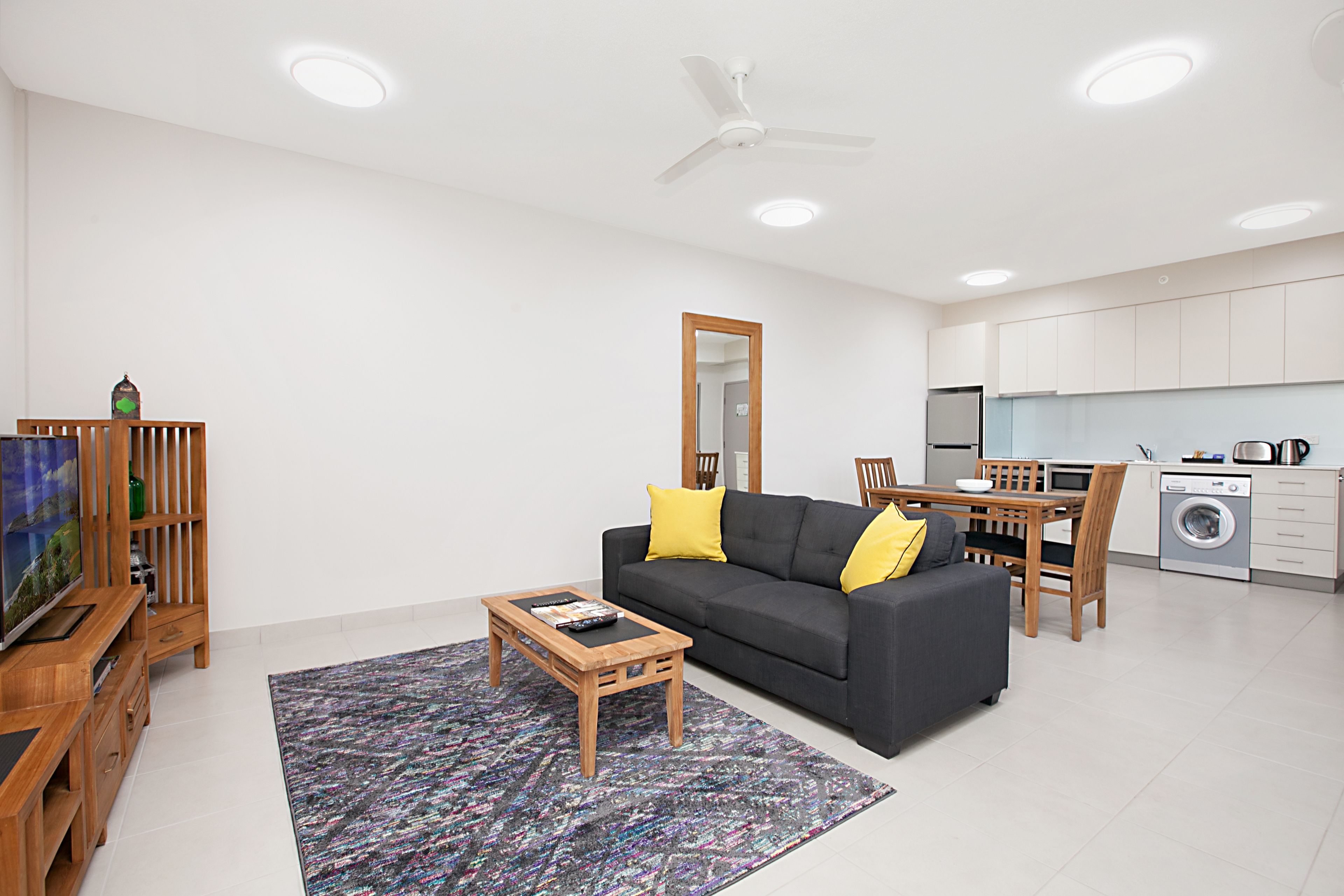A modern living room with a sofa, coffee table, and TV, a dining area with chairs, and a kitchen with appliances.