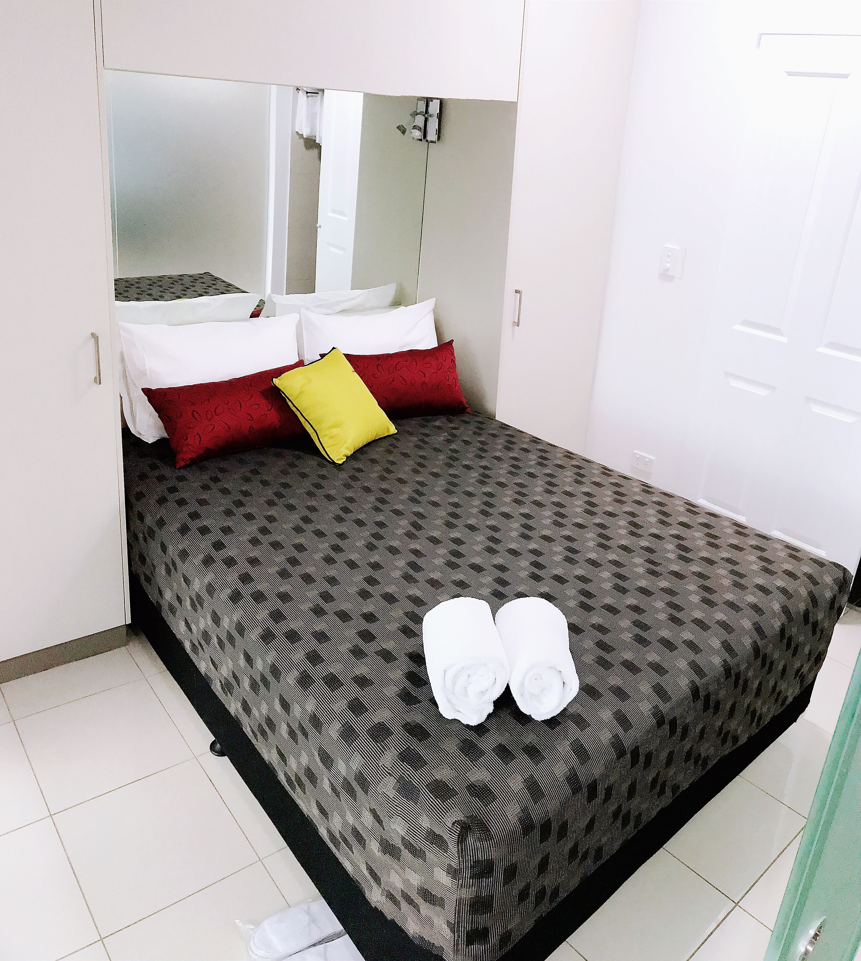 A neatly made bed with patterned bedding, a mirror above it, and a pair of white towels on the floor.