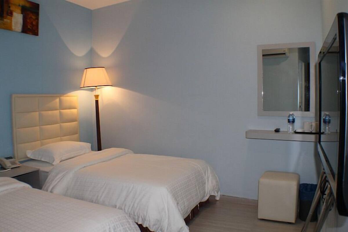 A hotel room with two beds, a bedside lamp, a small table with a phone, a mirror, and a bench.
