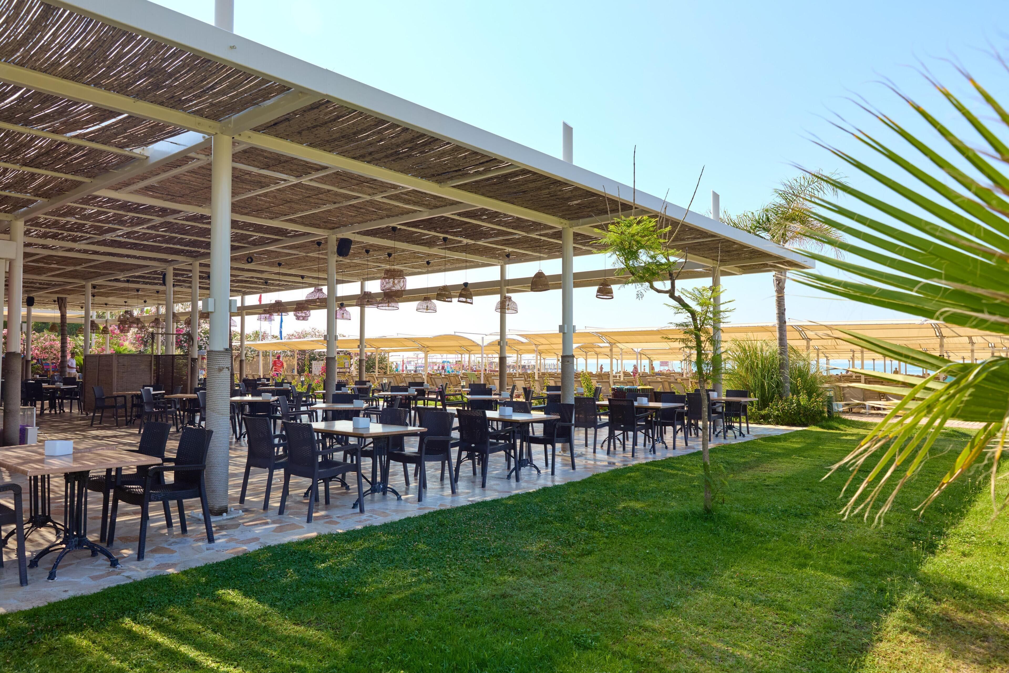 An outdoor dining area with tables and chairs, covered by a pergola, and surrounded by greenery.