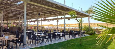 An outdoor dining area with tables and chairs, covered by a pergola, and surrounded by greenery.