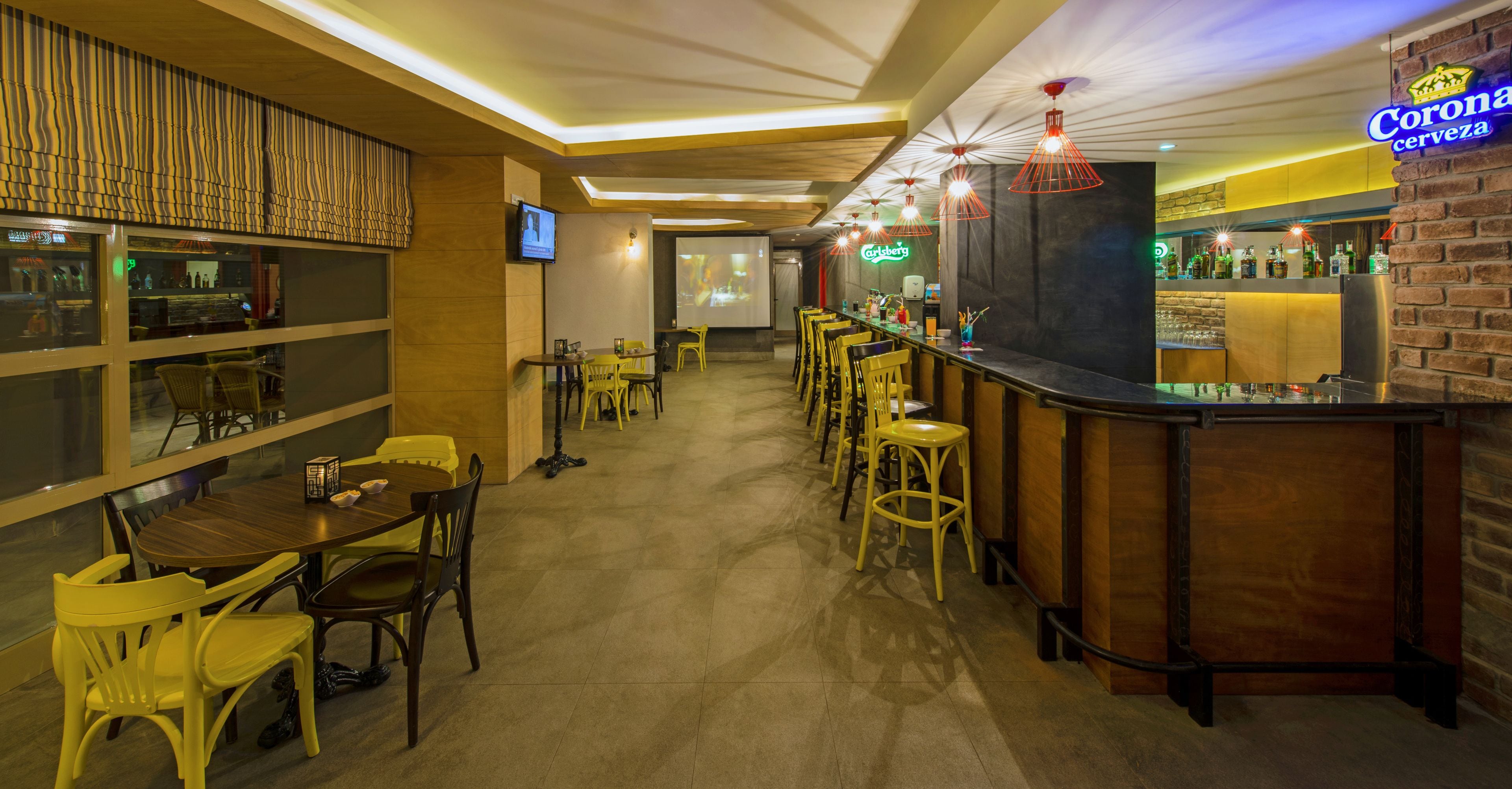 Uma área de bar com um anúncio da Corona, uma tela de projeção e diversos tipos de assento, incluindo banquetas e cadeiras.