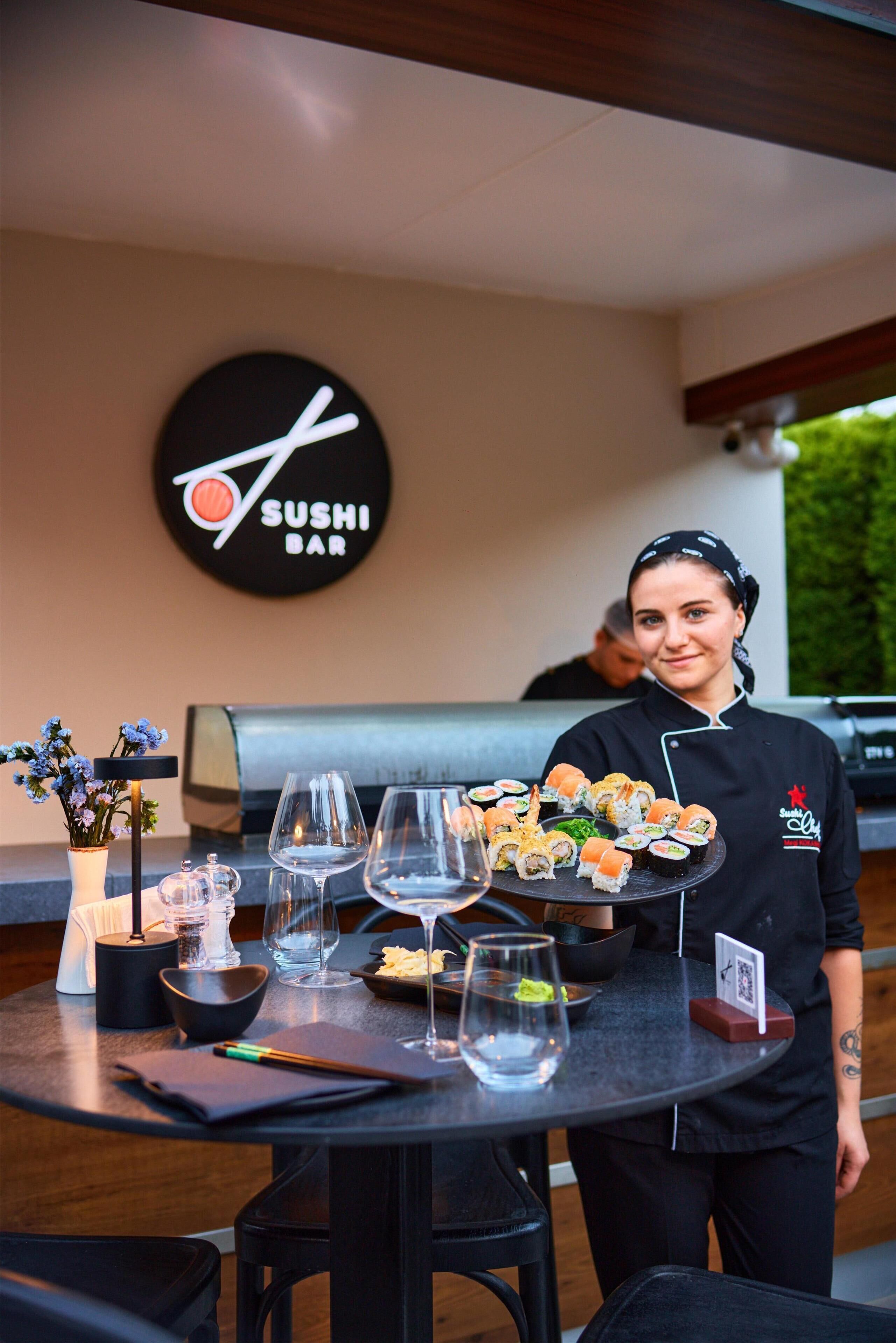 A sushi bar with a sign, a table with sushi, wine glasses, and a vase with flowers.