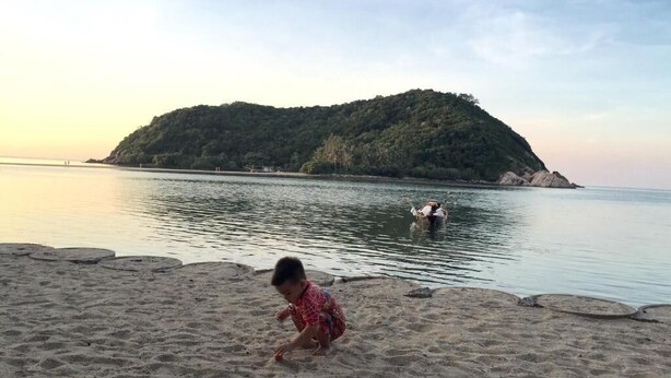 A beach with a child playing in the sand, a small boat in the water, and a forested island in the background. - Maehaad Guesthouse (Ko Pha-ngan)