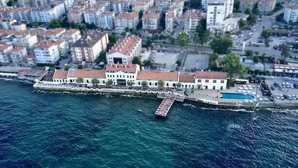 Vue aérienne d’une ville côtière, avec un imposant bâtiment blanc doté d’un toit rouge, une jetée s’avançant dans la mer et un espace piscine.
