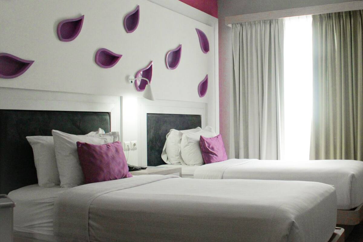 Ein Hotelzimmer mit zwei Betten, einem großen Fenster mit Vorhängen und einer Wand mit dekorativen, violetten Blattmustern.