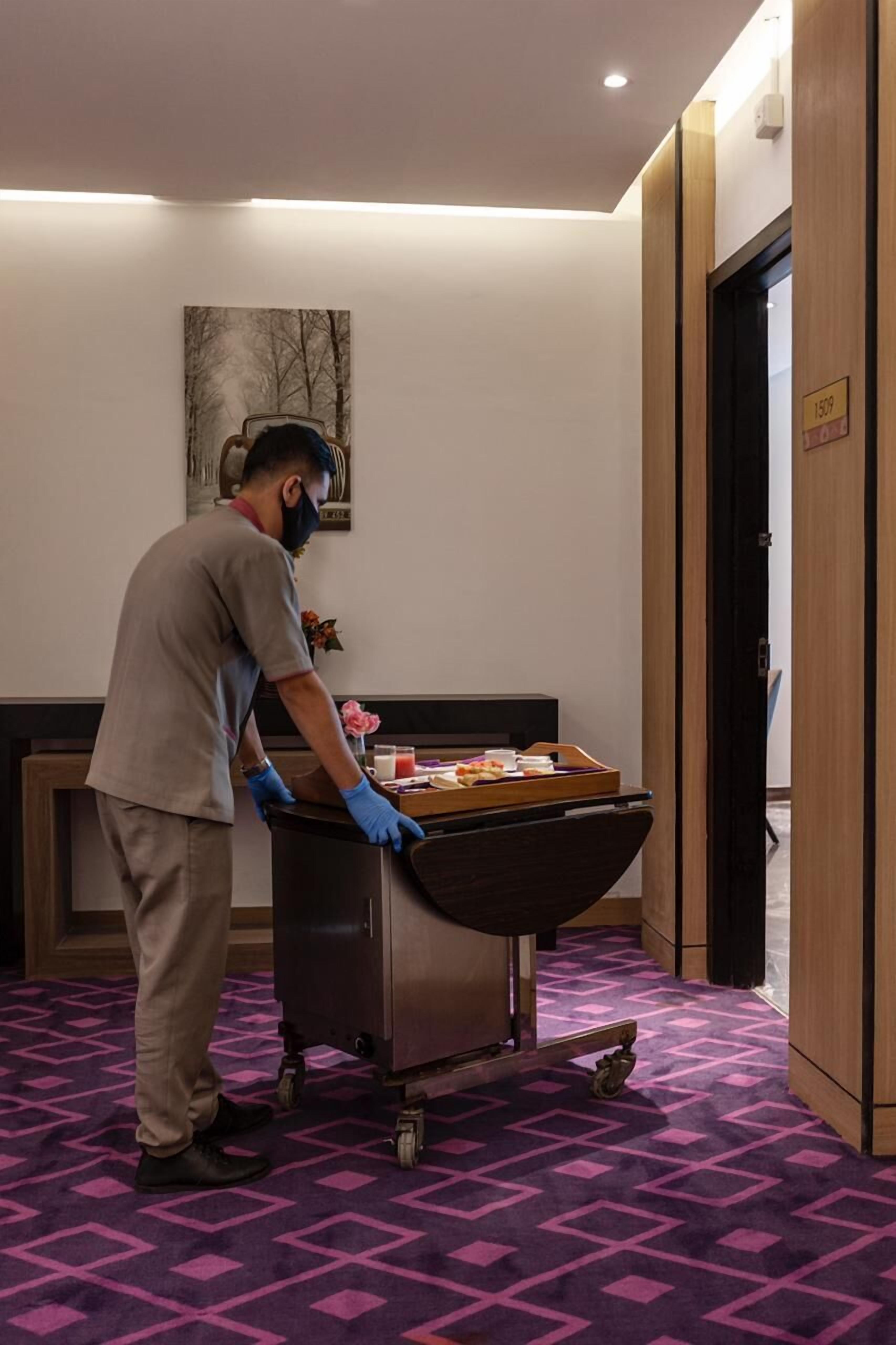 Een persoon in uniform maakt een kar schoon, compleet met een dienblad vol spullen, in een hotelkamer met een paars kamerbreed tapijt en een schilderij aan de muur.